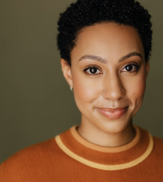 Close-up of a woman with short curly black hair, brown eyes, and light skin, smiling and wearing a burnt orange sweater with yellow and orange stripes around the collar, against a muted green background.