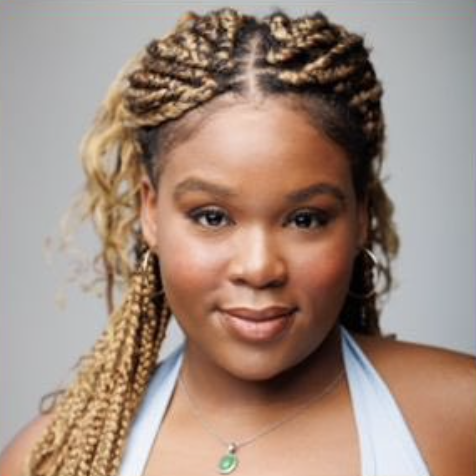 A woman with blonde and brown box braids wearing a blue top and a necklace with a green pendant.