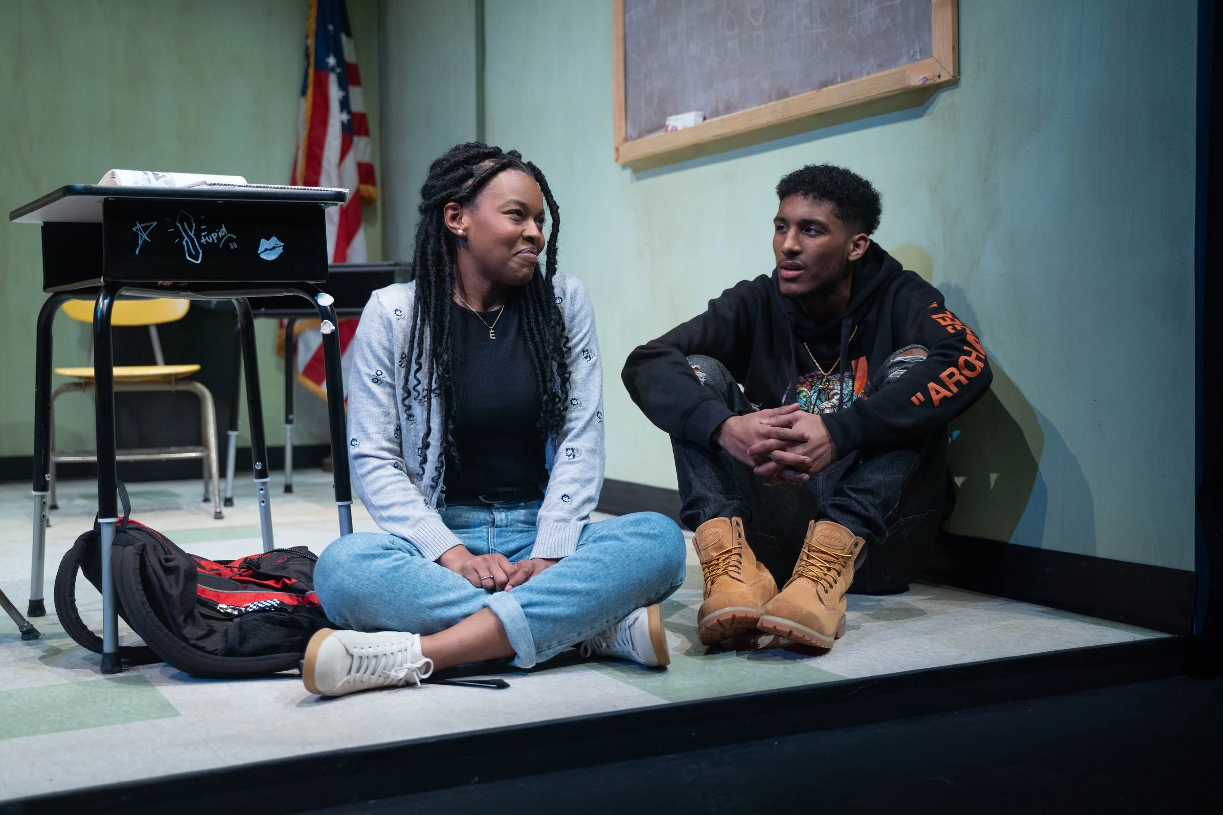 A young woman and a young man sitting on the floor of a classroom, engaged in a conversation. The woman has long, twisted hair, and the man has short, curly hair. The woman is wearing a black shirt, a gray cardigan, and blue jeans. The man is dressed in a black hoodie with orange lettering, ripped black jeans, and tan work boots. A backpack is on the floor beside them, and there is a black chalkboard with writing and drawings behind them. An American flag is visible in the background.