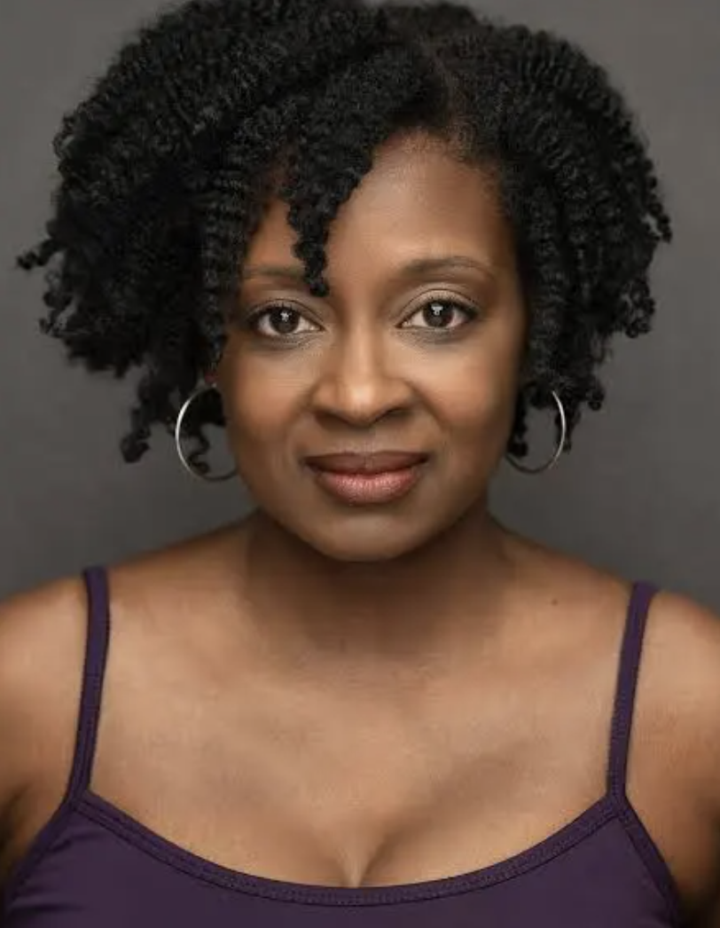 Close-up portrait of a woman with dark, curly hair wearing hoop earrings and a purple sleeveless top against a plain dark background.