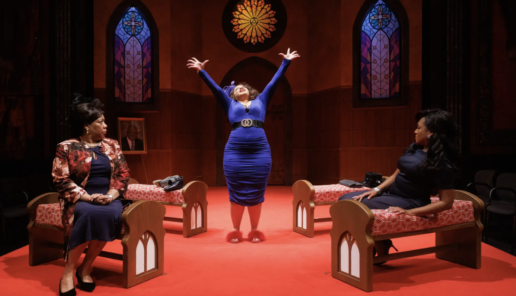 Three women in a church setting, with stained glass windows, two sitting on benches and one standing in the center with arms raised and in a bright blue dress. The woman on the left wears a floral jacket, and the woman on the right wears a dark dress. The scene appears theatrical.