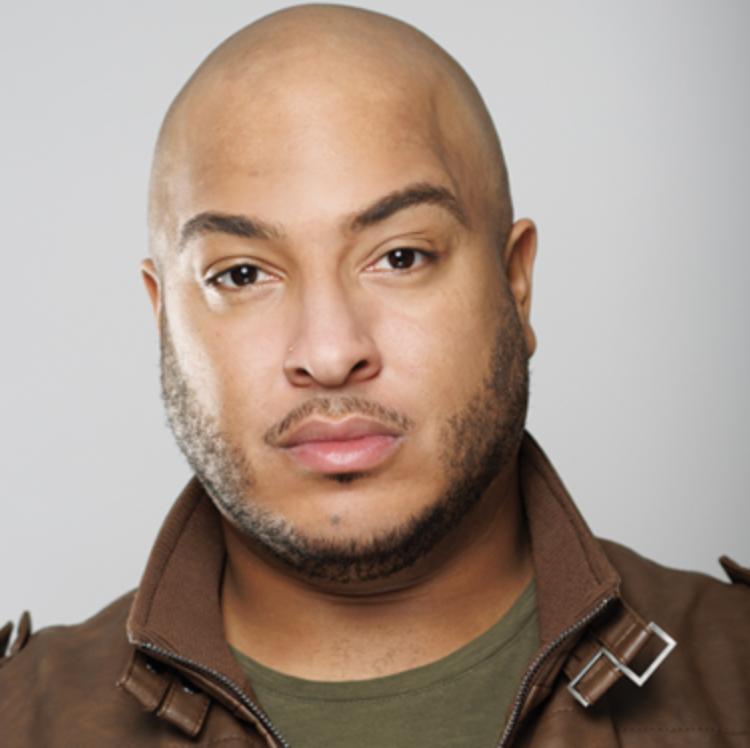 Close-up portrait of a man with a shaved head, light skin, and facial hair, wearing a brown jacket and an olive-green shirt against a plain light gray background.