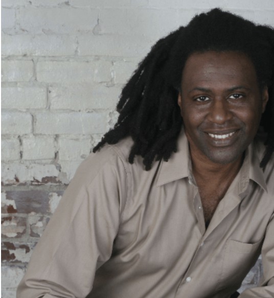 A man with long dreadlocks and dark skin, smiling, wearing a beige button-up shirt, against a white brick wall.