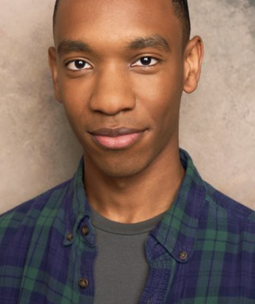 A young man with short dark hair and brown eyes, wearing a green and blue plaid shirt over a gray t-shirt, standing against a textured beige background.