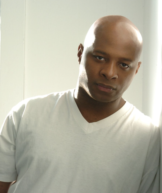 A bald man with a serious expression wearing a white t-shirt, standing by a window with sunlight coming through.