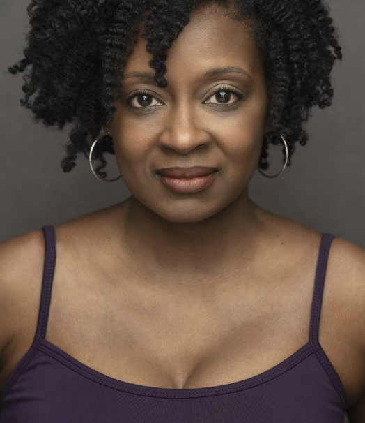 Close-up portrait of a woman with dark curly hair, wearing a purple tank top and hoop earrings, looking directly at the camera against a gray background.