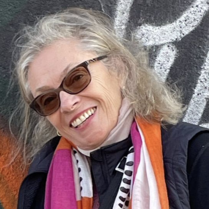 Smiling older woman with gray hair, wearing sunglasses, a black jacket, and colorful scarves, standing outdoors.