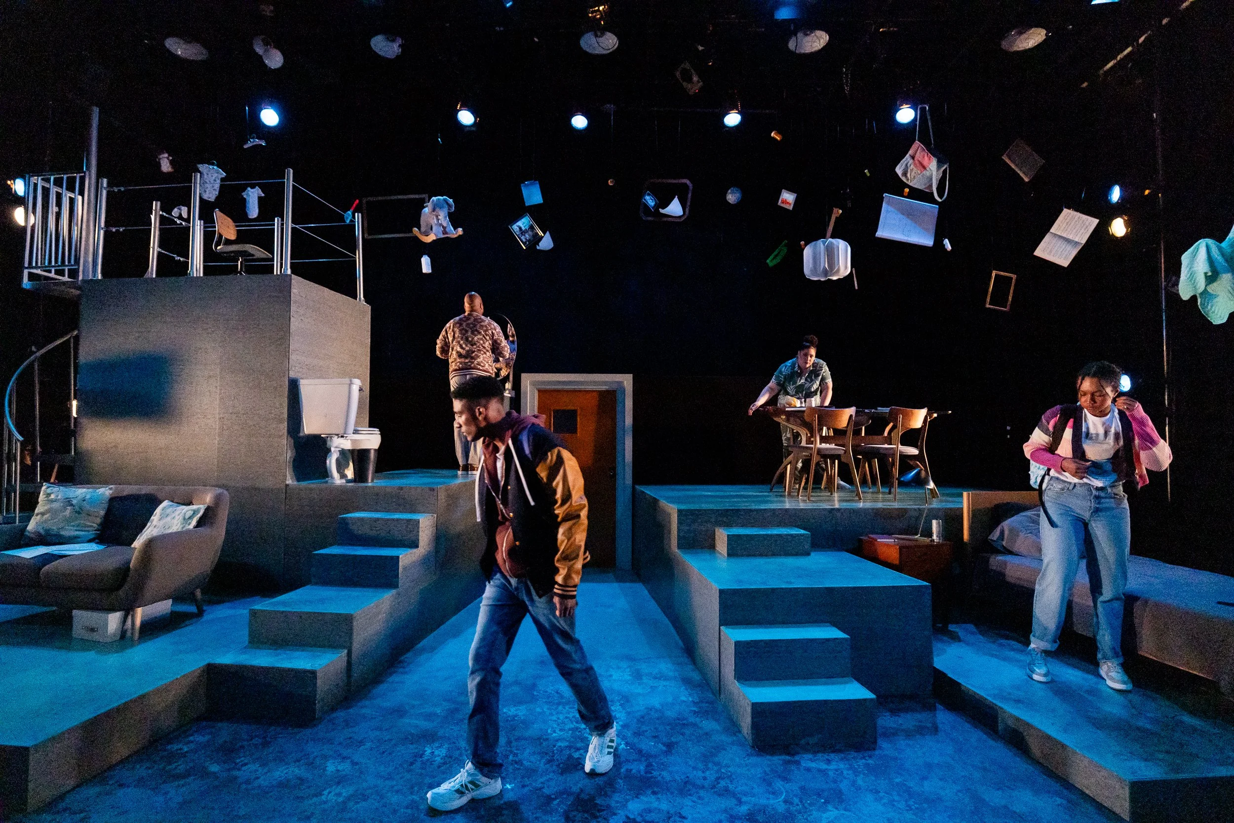 Stage set with hanging props, including furniture, books, and clothing, with three people preparing for a performance, lighting focused on the scene, and a cozy seating area in front.