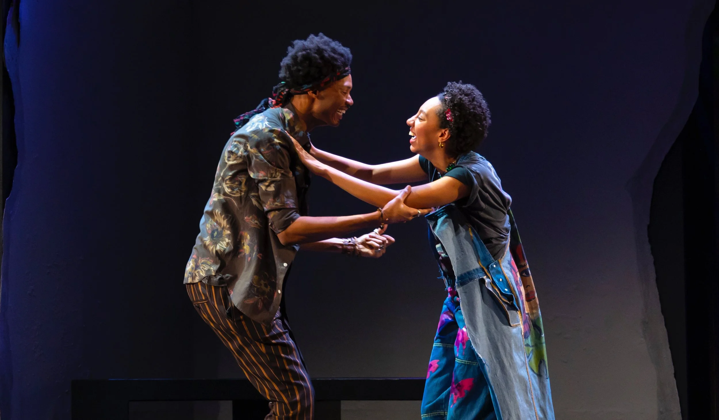 Two people, a man and a woman, smiling and holding each other's hands, standing close together on a stage with a dark background.