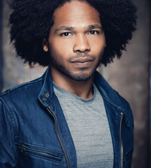 Close-up portrait of an African American man with a serious expression, wearing a denim jacket over a gray t-shirt.