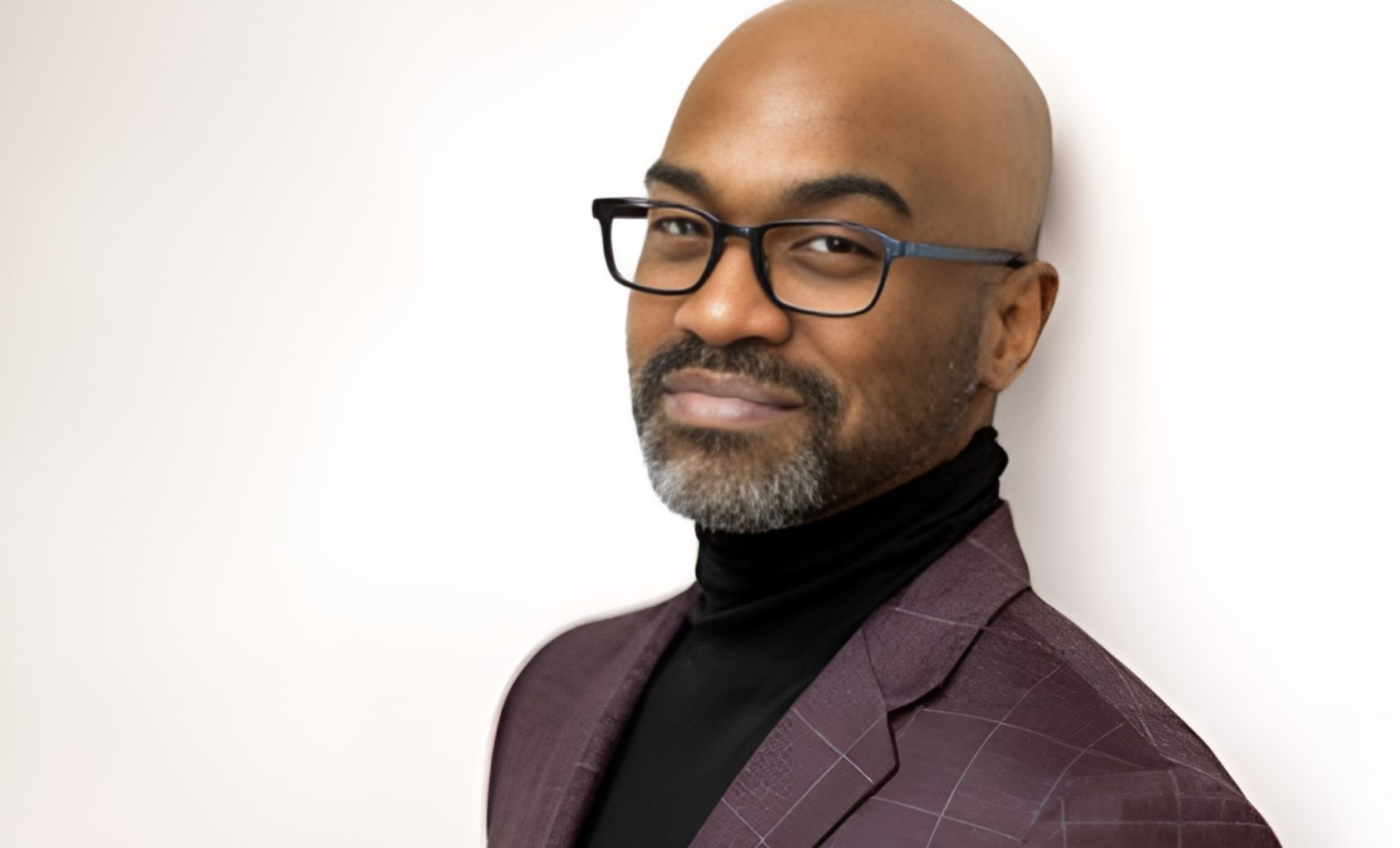 A man with glasses, a beard, and a shaved head, wearing a purple checkered suit jacket and a black turtleneck, standing against a plain white wall.