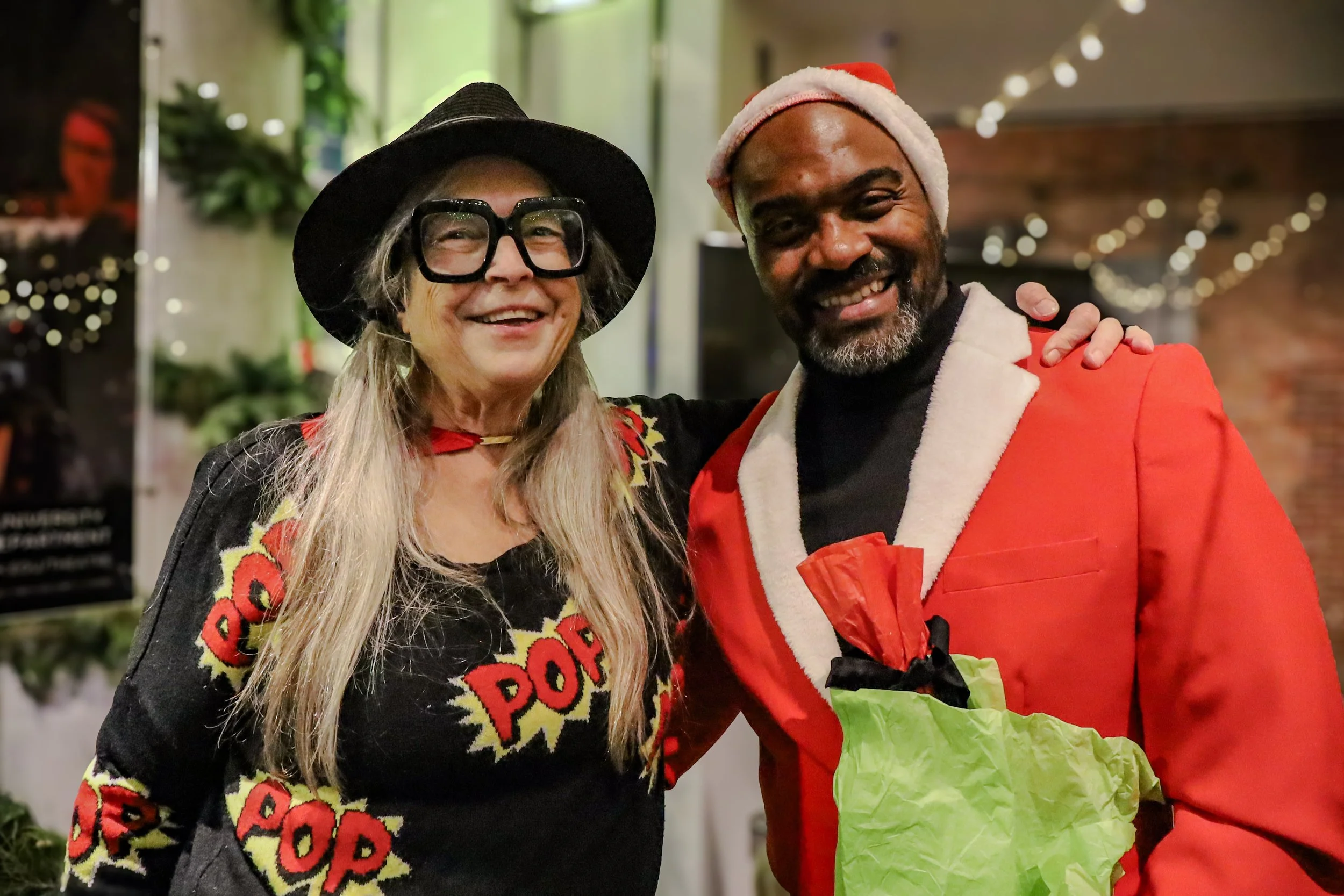 Two people smiling, dressed in festive holiday attire, one in a black hat and large glasses wearing a sweater with a pop art style design, and the other in a Santa hat and red jacket, holding a green paper bag.