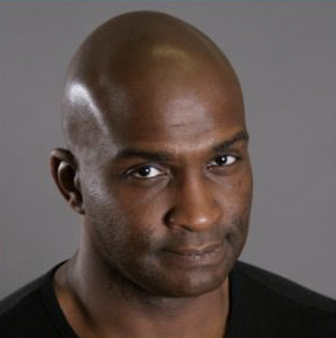 Close-up portrait of a bald man with a confident expression, wearing a black shirt, against a plain gray background.