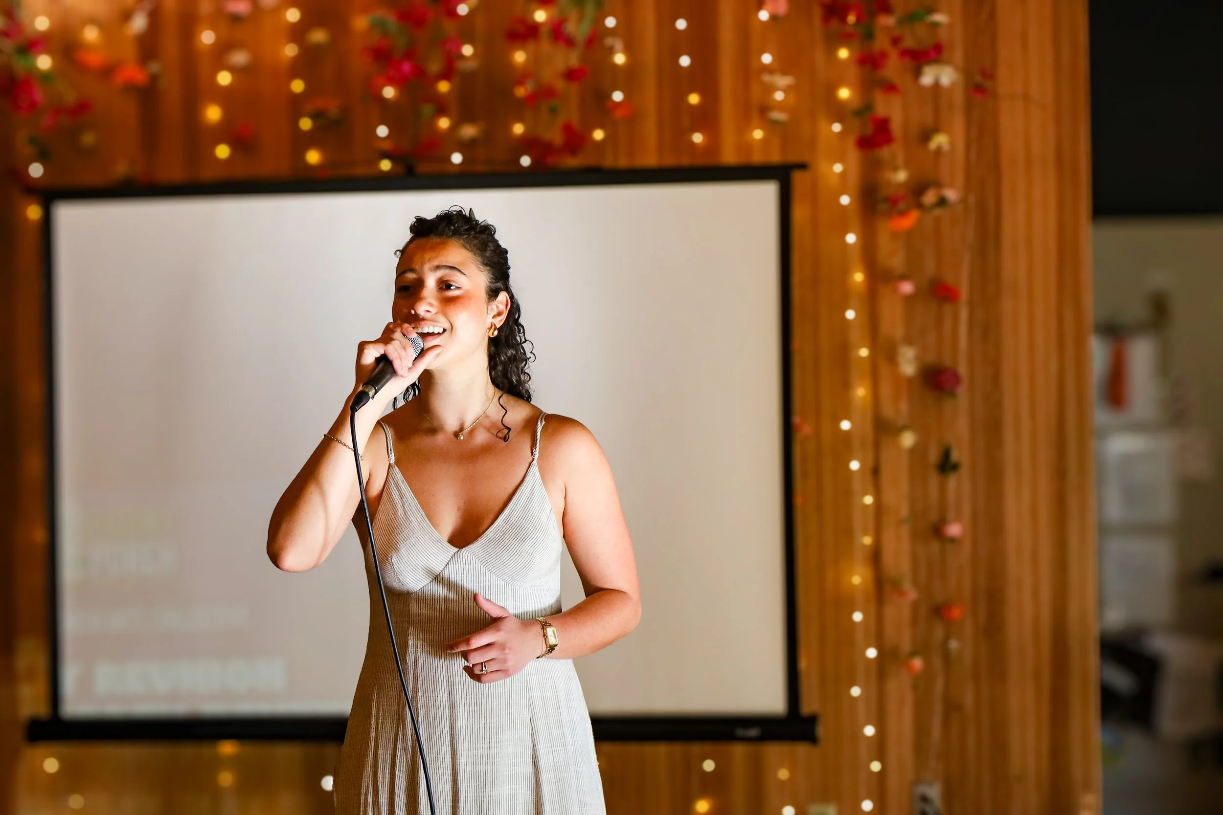 A woman in a beige dress holding a microphone, speaking at an indoor event with a wooden wall decorated with string lights and red and pink flowers in the background.