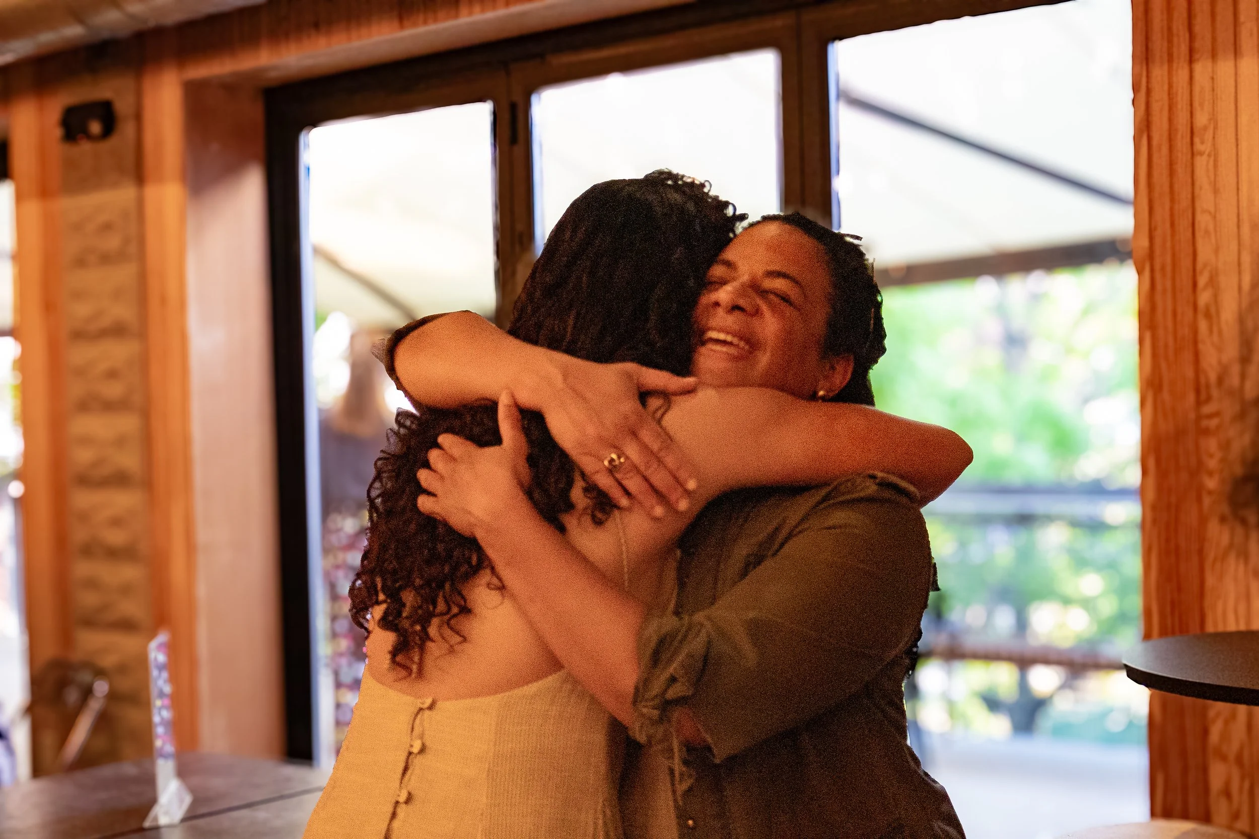 Two women hugging each other warmly in a cozy indoor setting with wooden walls and windows in the background.