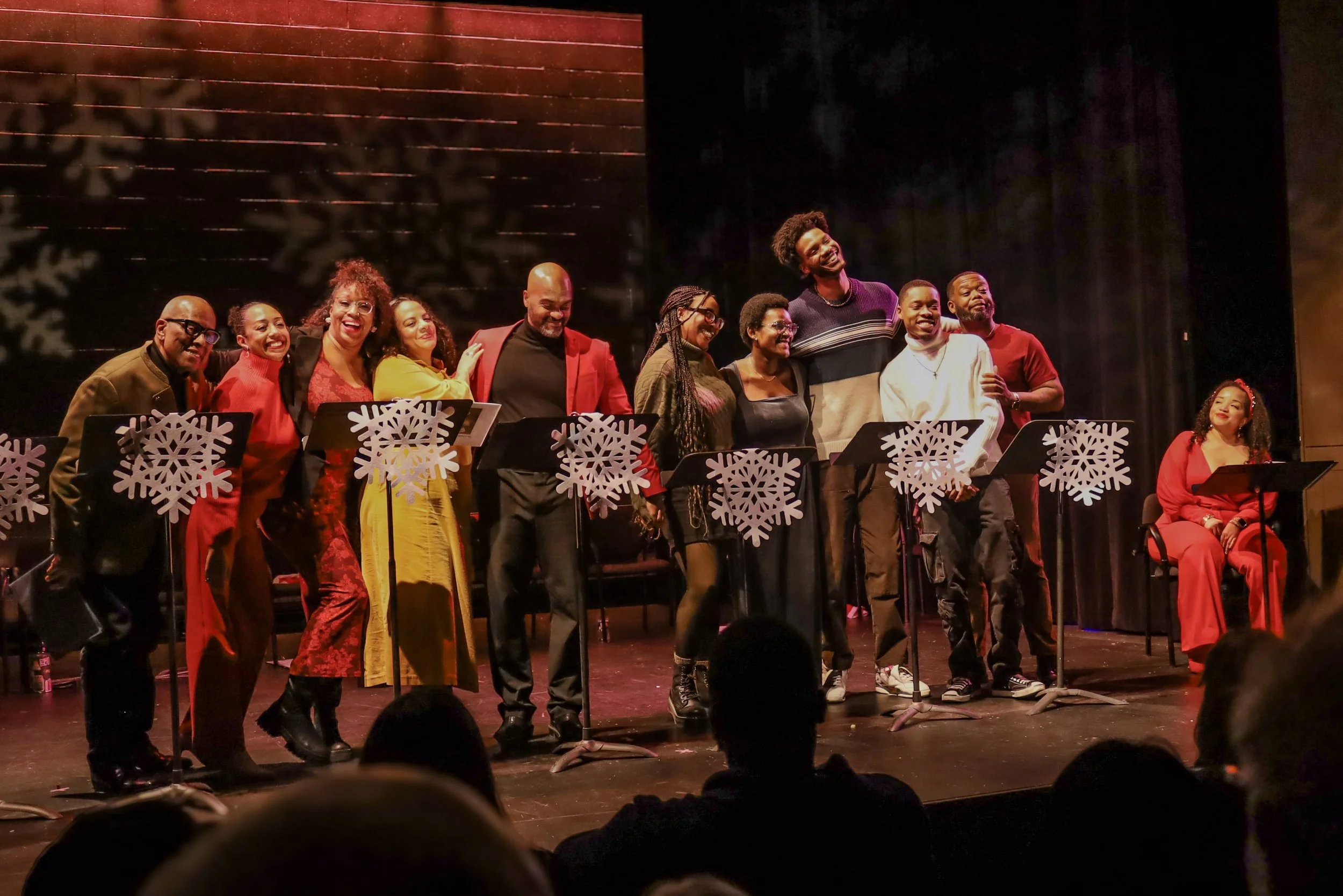 A group of diverse performers on stage, dressed in festive attire, smiling and embracing after a performance. They are standing behind music stands decorated with snowflakes, indicating a holiday or Christmas concert.