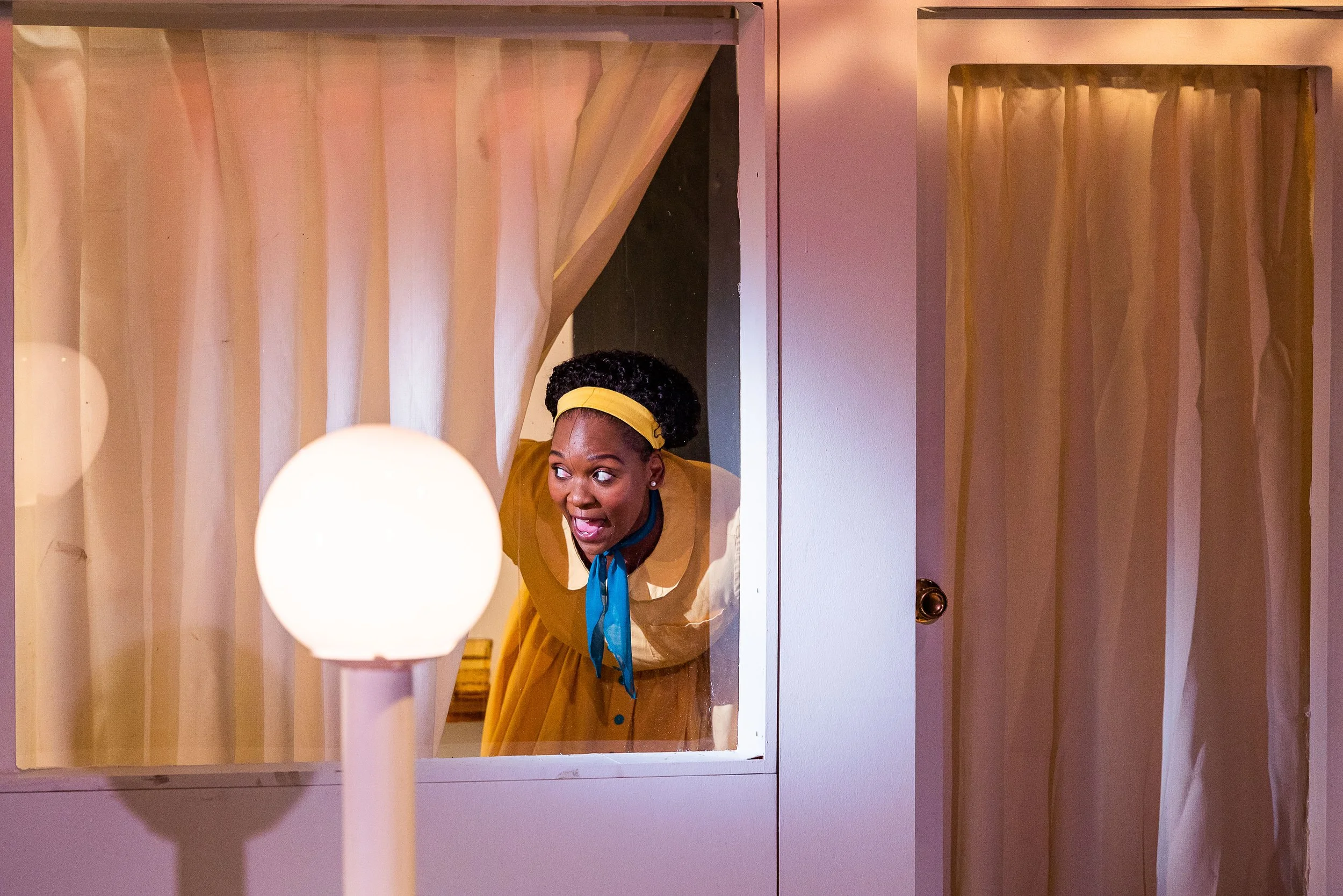 Woman wearing a yellow dress and headband looking through a mirror with wooden framing, with a glowing round light in the foreground and a closet door on the right.