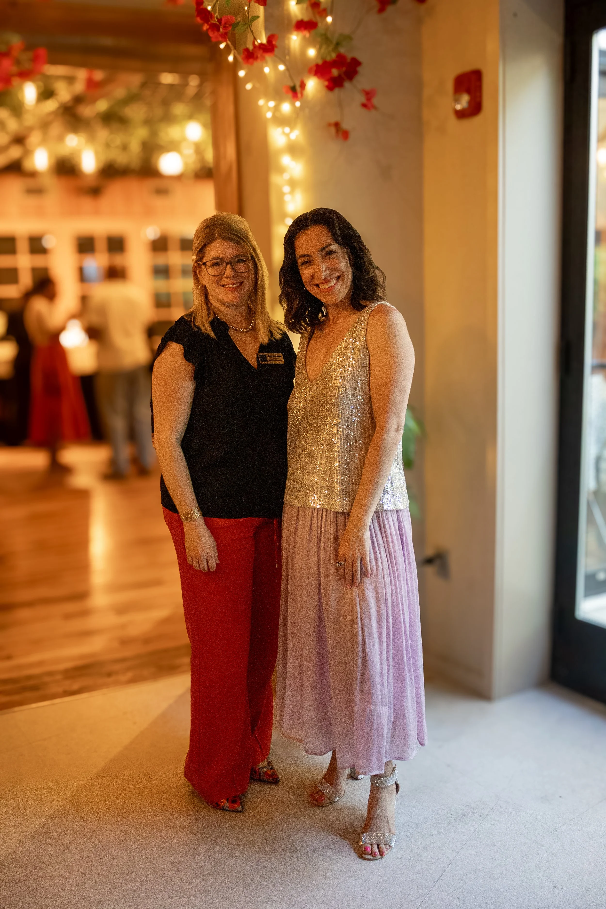 Two women standing together indoors, smiling at the camera. The woman on the left has blonde hair, glasses, and is wearing a black top with red pants. The woman on the right has dark wavy hair and is wearing a sparkly gold sleeveless top with a pink 