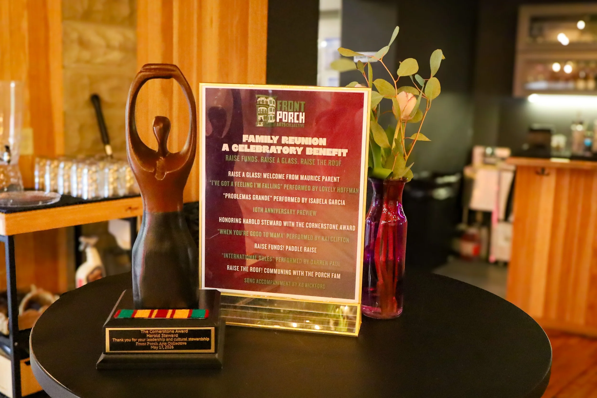 Award display on a round black table featuring a sculpture, a framed program, and a plaque; the sculpture is wooden with an abstract design, the framed program listing event details and performers, and the plaque thanks Harold Steward for leadership 