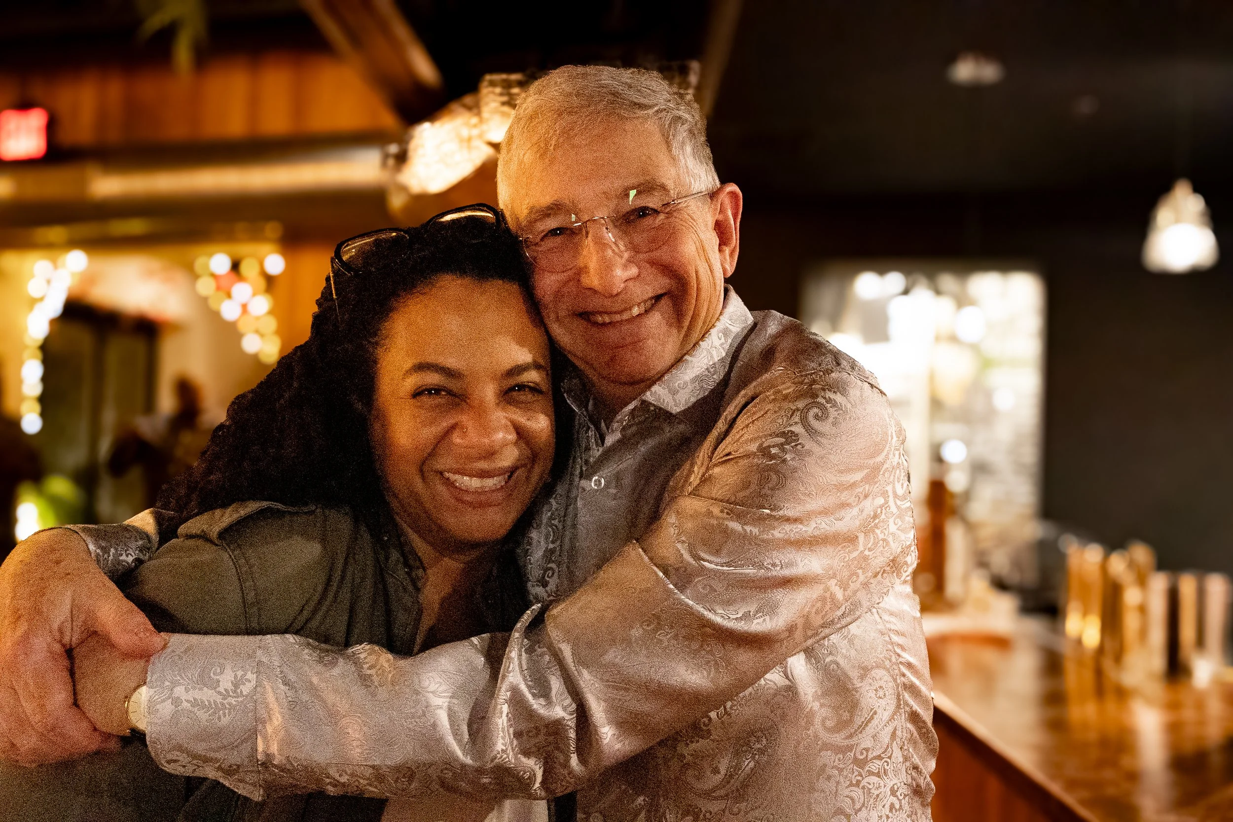 Two people smiling and hugging in a warmly lit indoor setting with wooden accents and blurred background.