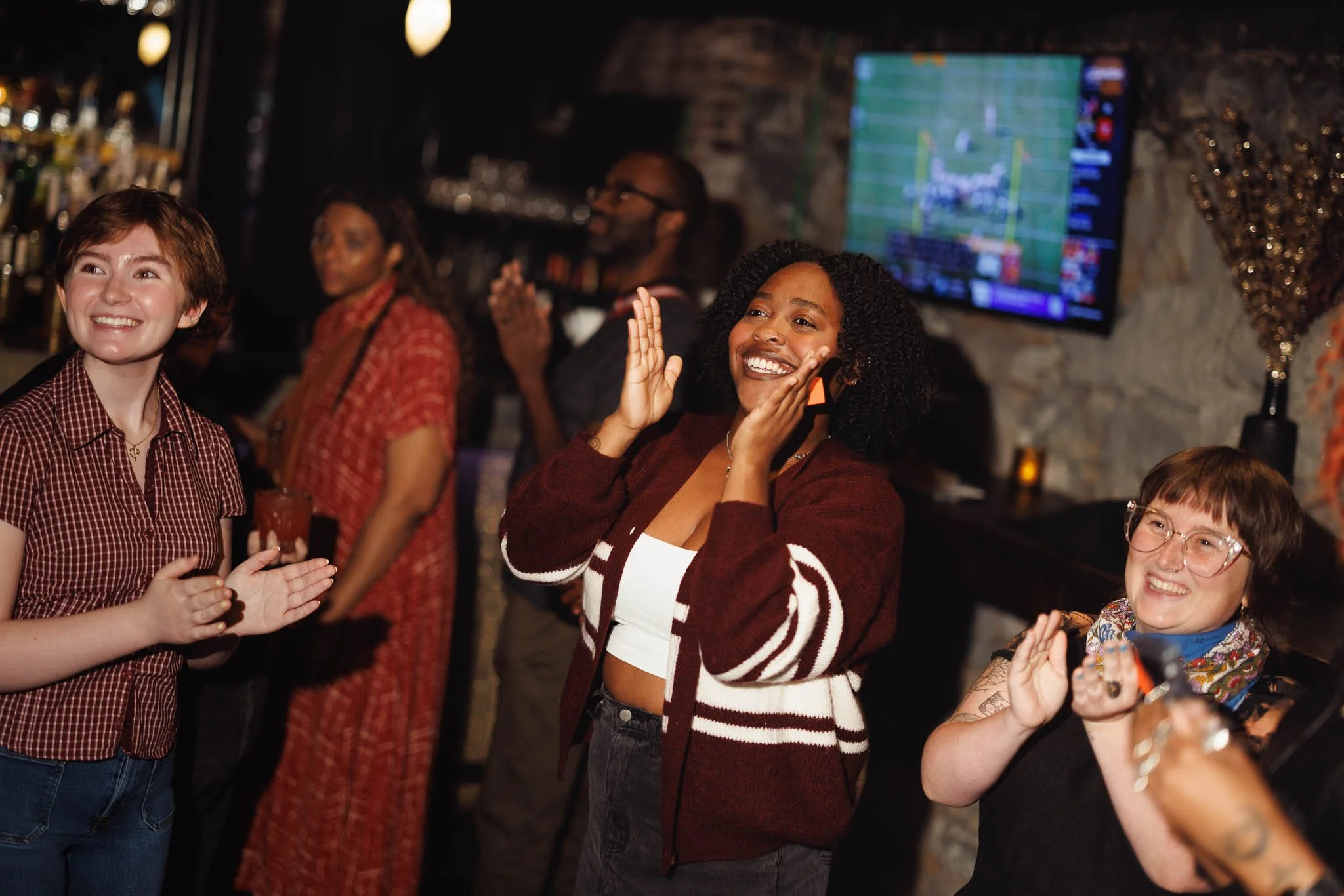 Group of smiling people at a social gathering, with a woman in the center clapping and enjoying the moment, in a dimly lit bar or restaurant setting.