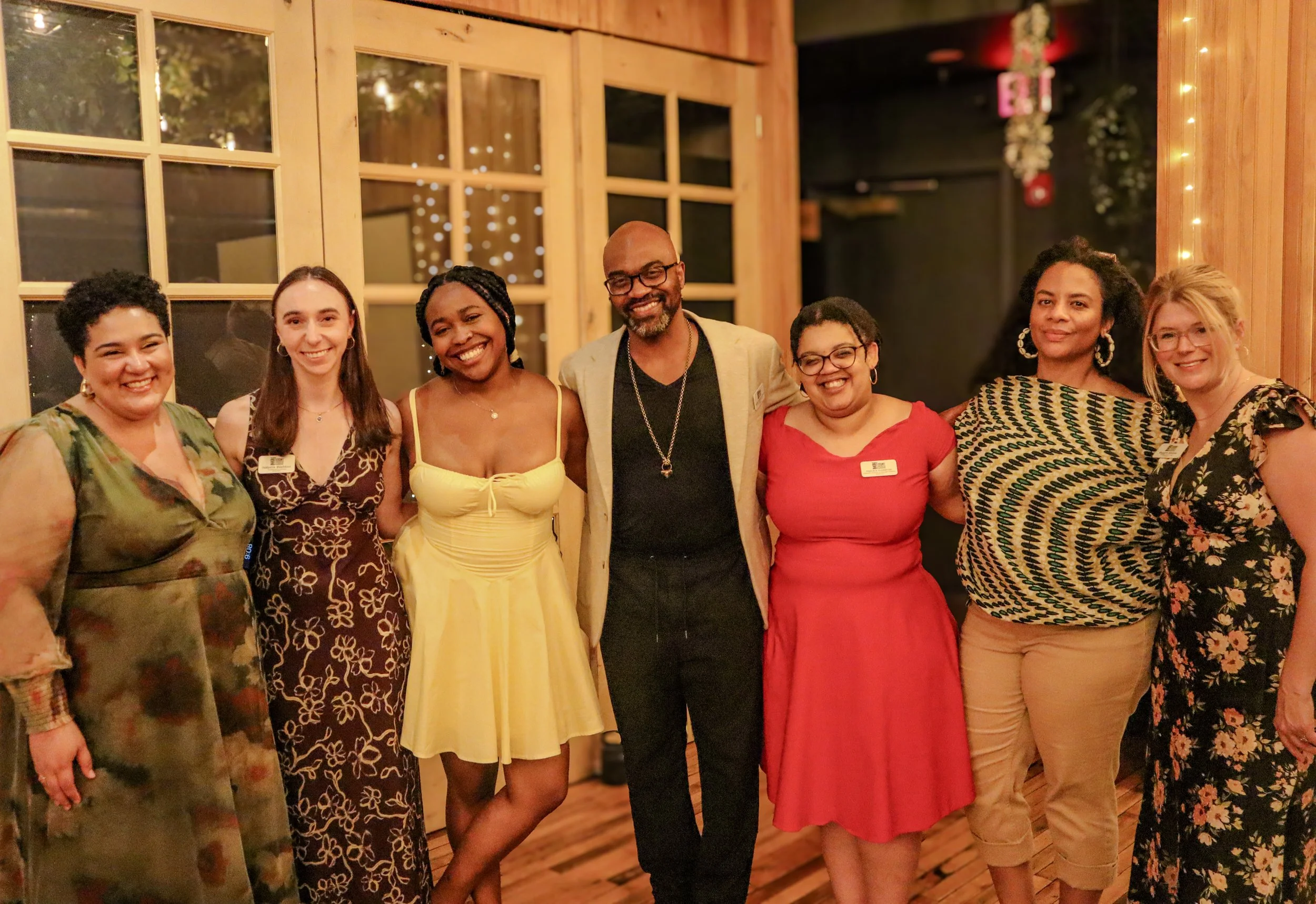 Group of seven diverse women and one man standing together indoors at a social event, smiling and posing for the camera.
