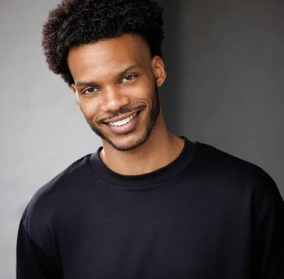 Smiling young man with curly hair, wearing a black shirt, standing against a gray and dark background.