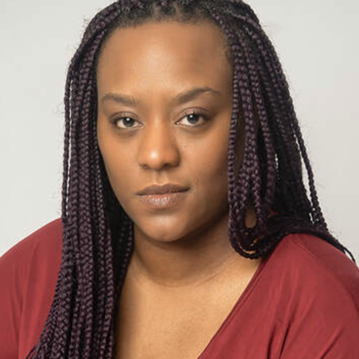 A young woman with long braided hair wearing a red top, looking directly at the camera with a neutral expression.