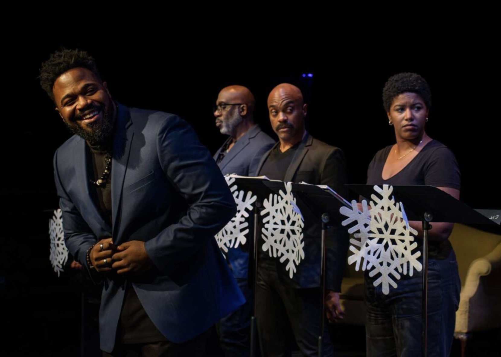 Four African American people standing on stage with music stands decorated with snowflakes, two men and one woman looking serious and one man smiling, dressed in black or dark clothing, under dark lighting.