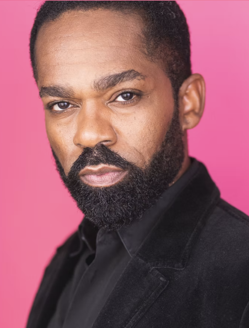 Close-up portrait of a Black man with a trimmed beard and short hair, wearing a black jacket, against a pink background.