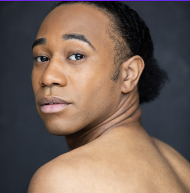 Close-up portrait of a young Black man with braided hair and bare shoulders against a dark background.