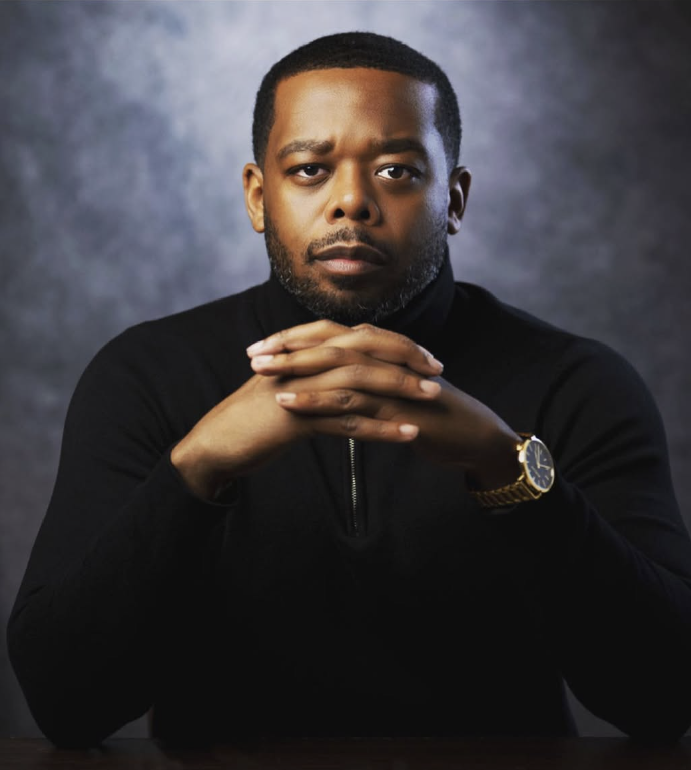 A man with a serious expression, wearing a black long-sleeve shirt and a gold watch, with hands clasped together, against a dark gray background.