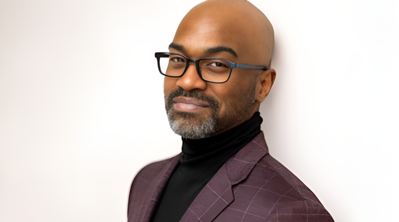 A bald man with glasses, a beard, and a mustache, smiling slightly while looking at the camera, wearing a checkered maroon blazer and a black turtleneck, standing against a white background.