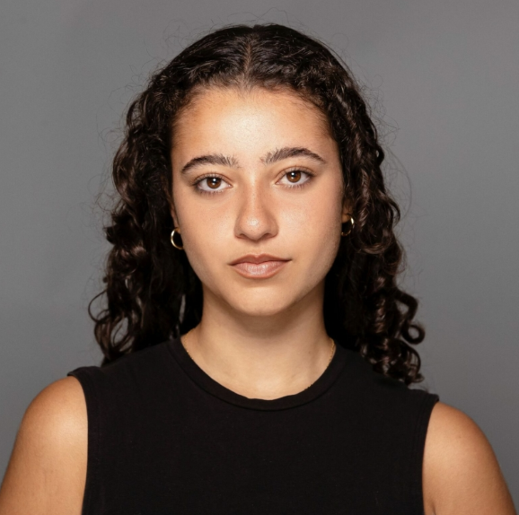 A young woman with dark, curly hair wearing a sleeveless black top and gold hoop earrings, looking directly at the camera against a plain gray background.