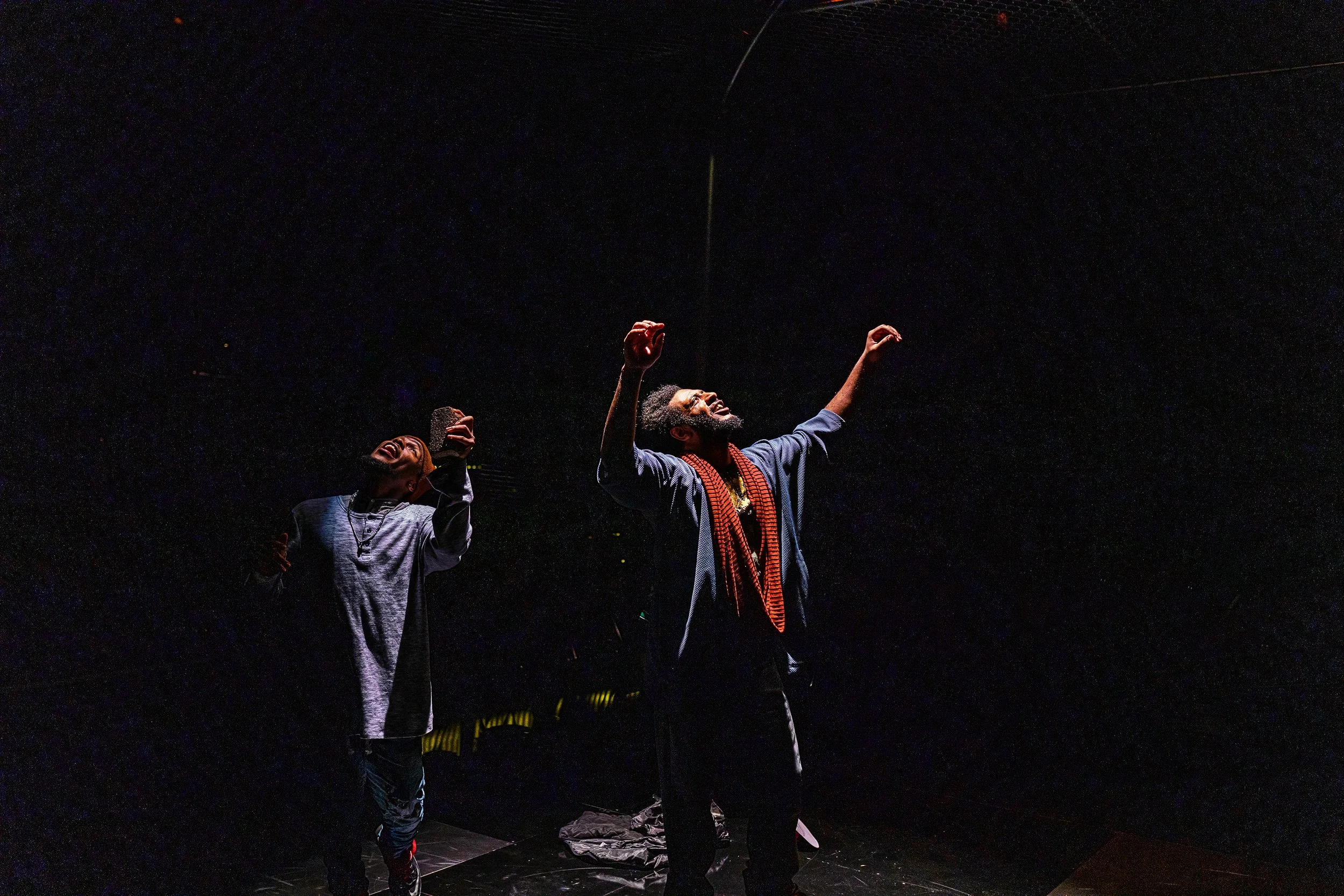 Two men are on stage under a dark spotlight, with their arms raised and joyful expressions. The man on the right is dressed in a dark blazer with a colorful scarf, and the man on the left is in a light-colored shirt. The background is dark with faint hints of stage equipment.