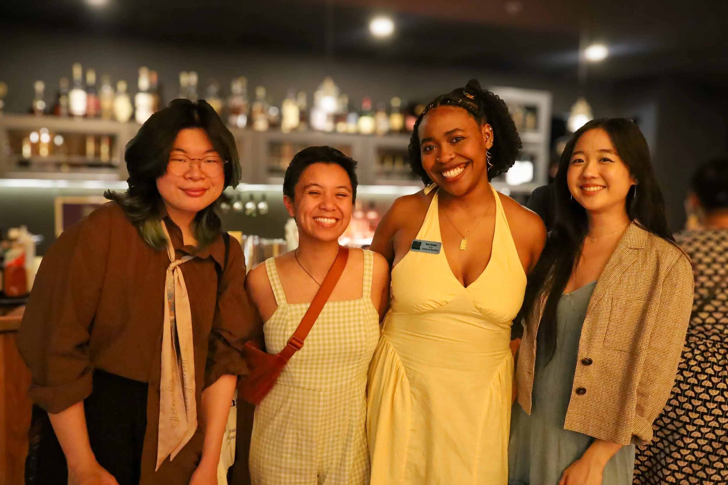 Four women smiling in a bar or restaurant setting, with bottles of alcohol displayed on shelves behind them.