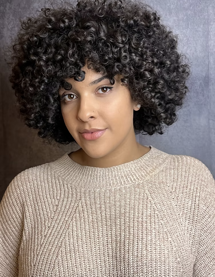 A young woman with brown skin and short, curly black hair, wearing a beige knit sweater and pearl earrings.