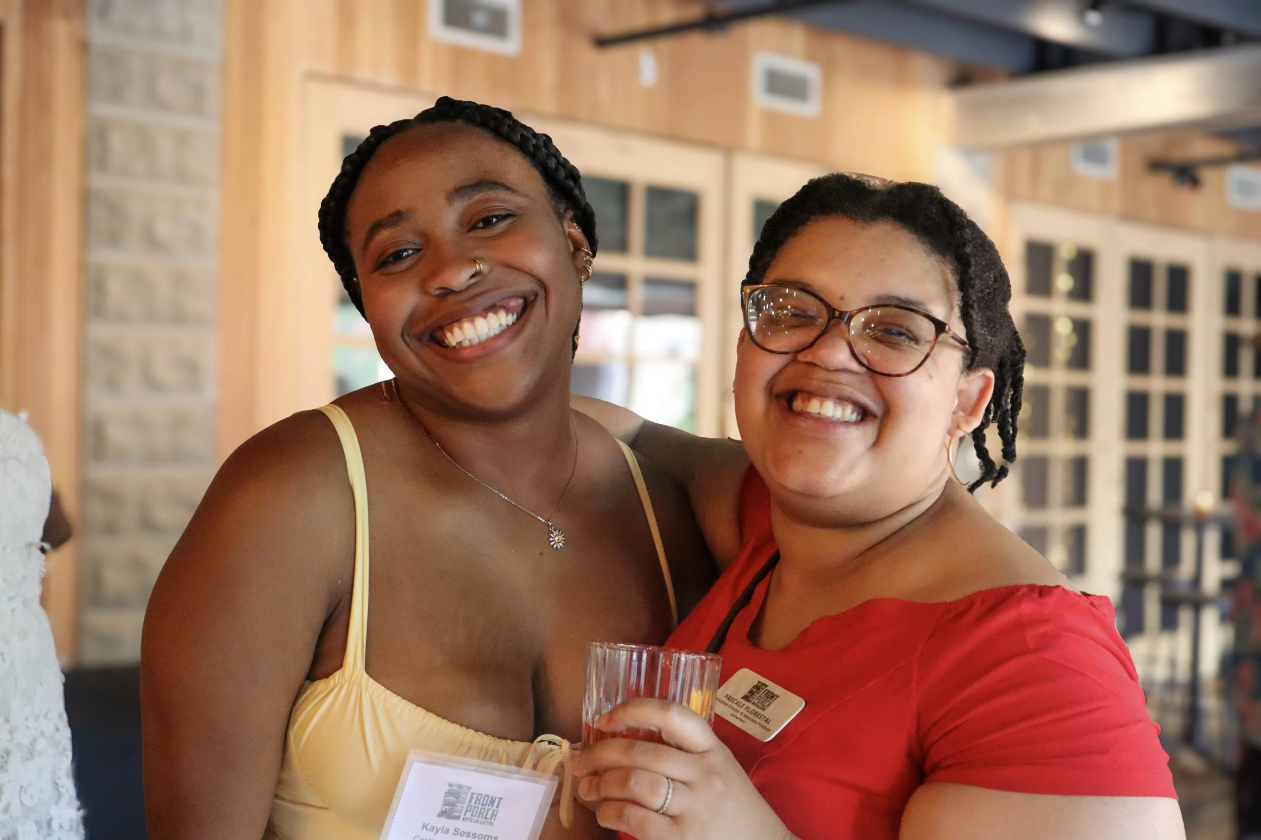 Two women smiling and hugging at an indoor event. One woman has dark skin, braided hair, and is wearing a yellow dress; the other woman has light skin, glasses, and is wearing a red top. They are holding a glass and have name tags on their chests.