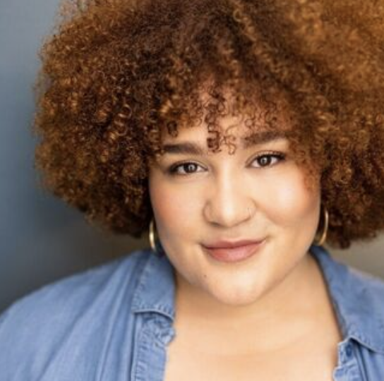 A woman with curly, reddish-brown hair smiling at the camera, wearing hoop earrings and a denim shirt.