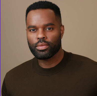 Portrait of a serious-looking Black man with short hair and a beard, wearing a dark shirt, against a beige background.