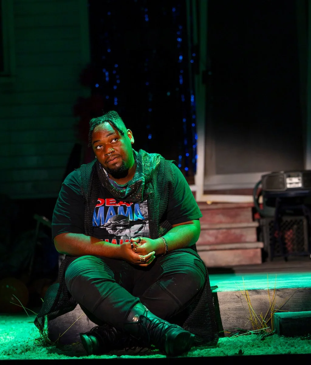A man sitting cross-legged outdoors at night, illuminated by green stage lights, with a confused or thoughtful expression, in front of a wooden house with a door and a crate.