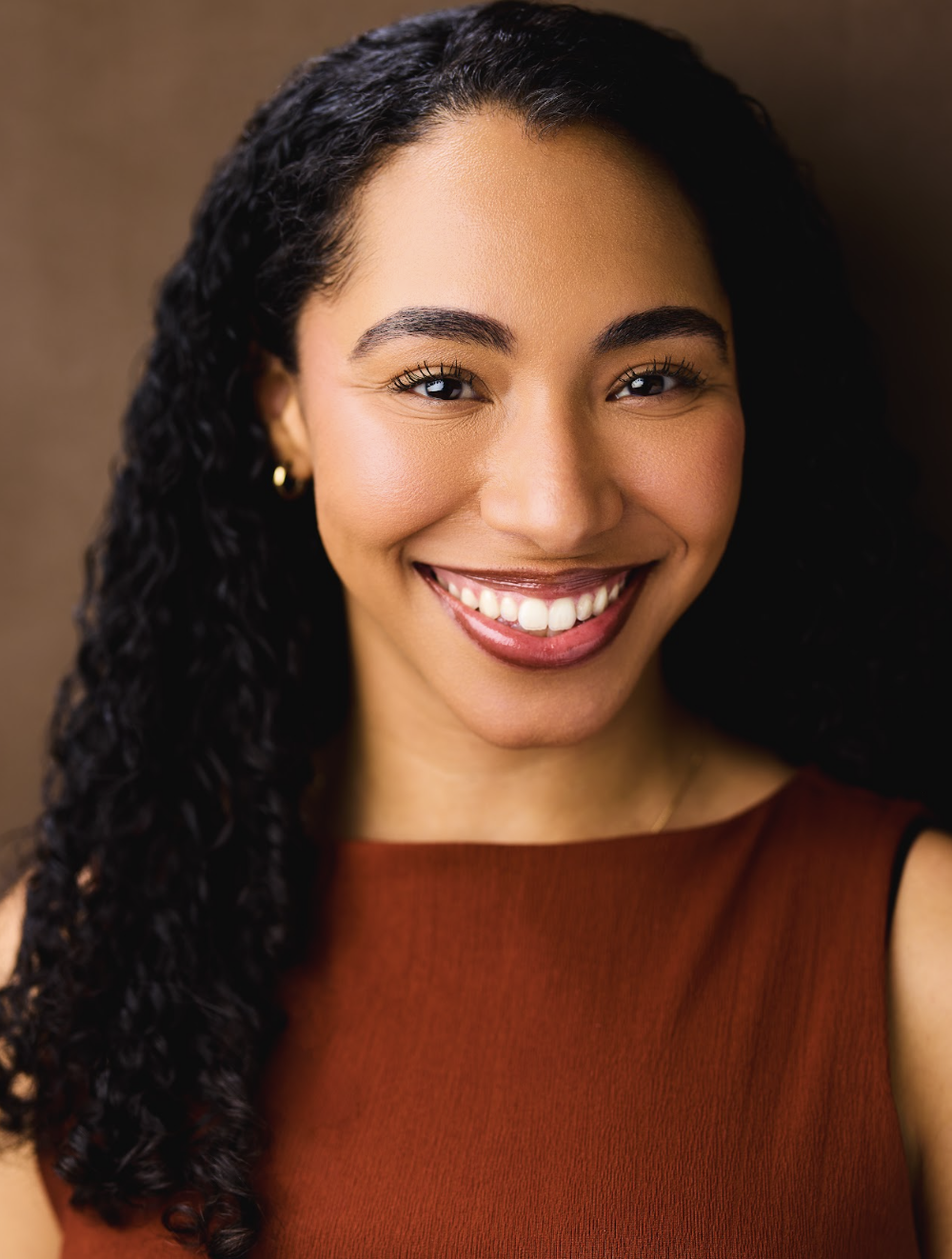 Close-up portrait of a smiling woman with curly black hair, wearing a sleeveless maroon top and gold hoop earrings, against a brown background.