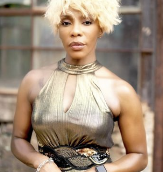 Woman with blonde curly hair wearing a metallic sleeveless top with a keyhole neckline, standing in front of a rustic background.