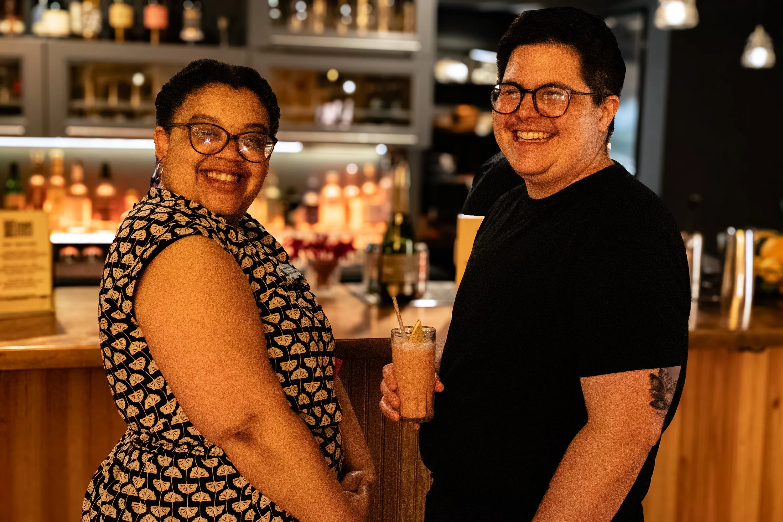 Two people, a woman and a man, smiling and holding a drink together at a bar or restaurant.