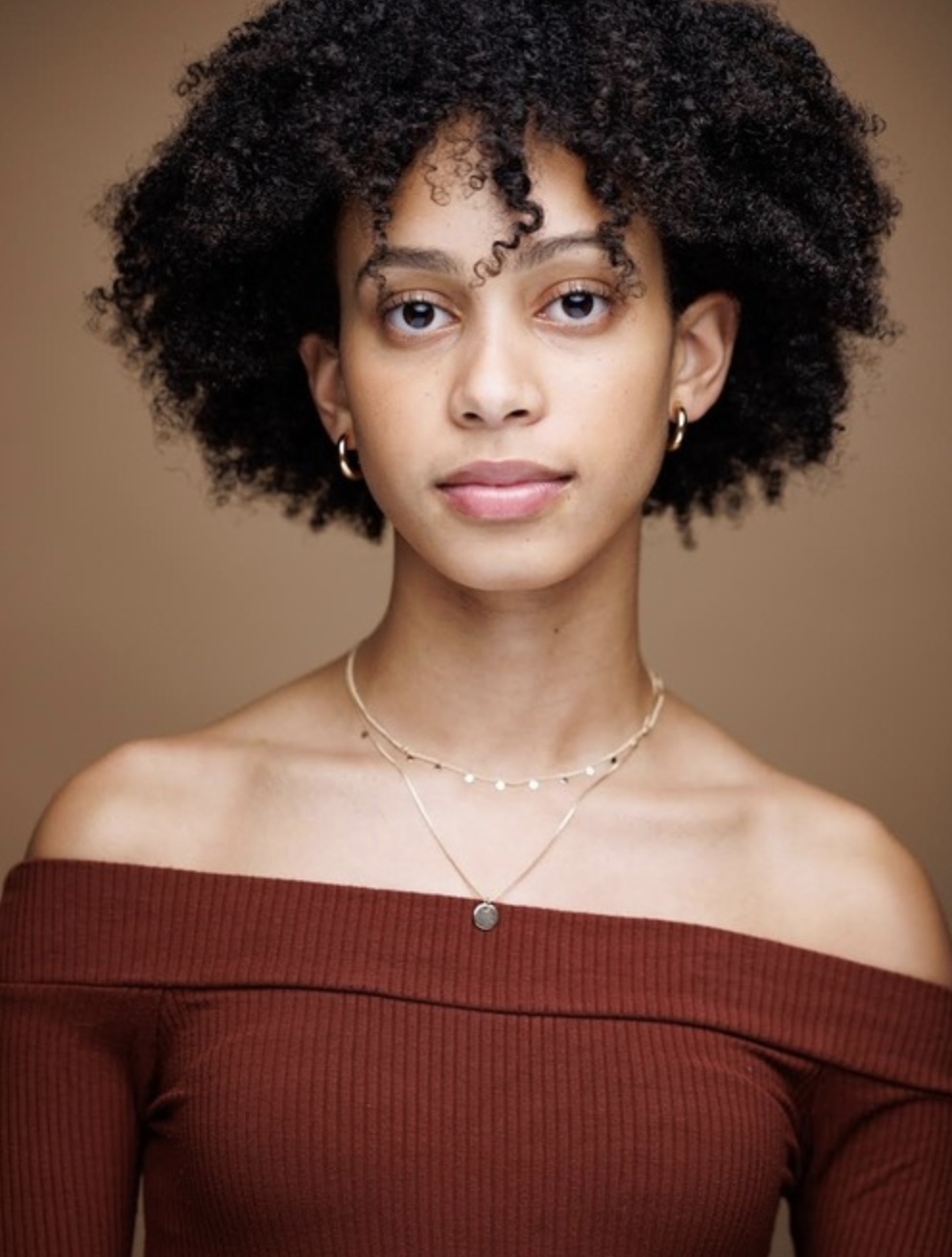 Portrait of a young woman with curly hair, wearing gold hoop earrings, layered necklaces, and an off-the-shoulder, ribbed, maroon top against a neutral background.