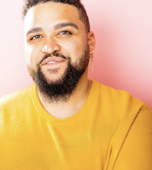 Portrait of a man with curly hair, beard, and mustache, wearing a yellow sweater and a gold earring, smiling slightly against a pink background.