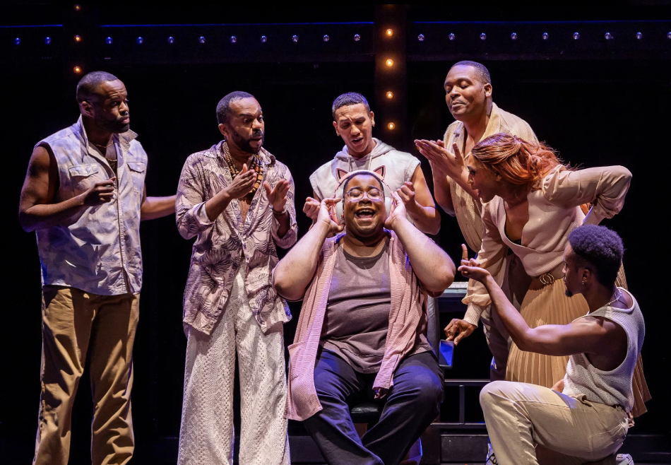 A group of seven diverse actors performing a theatrical scene on stage, with some clapping and others engaged in expressive gestures.
