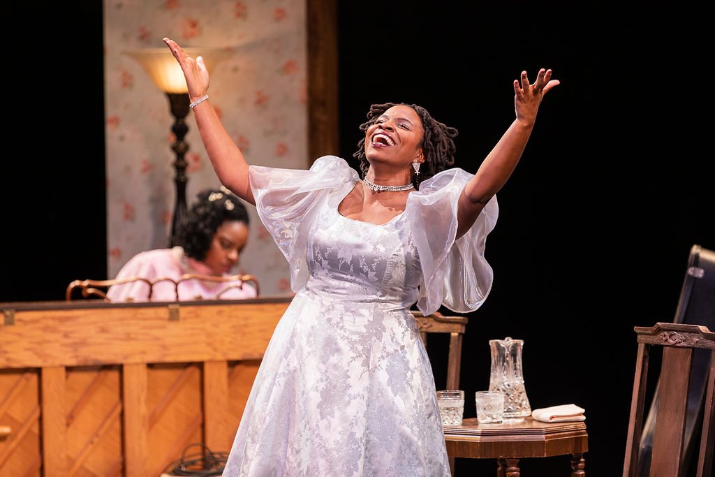 A woman in a white dress with puffy sleeves is joyfully singing or performing on stage with her arms raised. Another woman is seated at a piano in the background, with a table holding a glass pitcher and glasses in front of her. The scene appears to be a theatrical performance or concert.