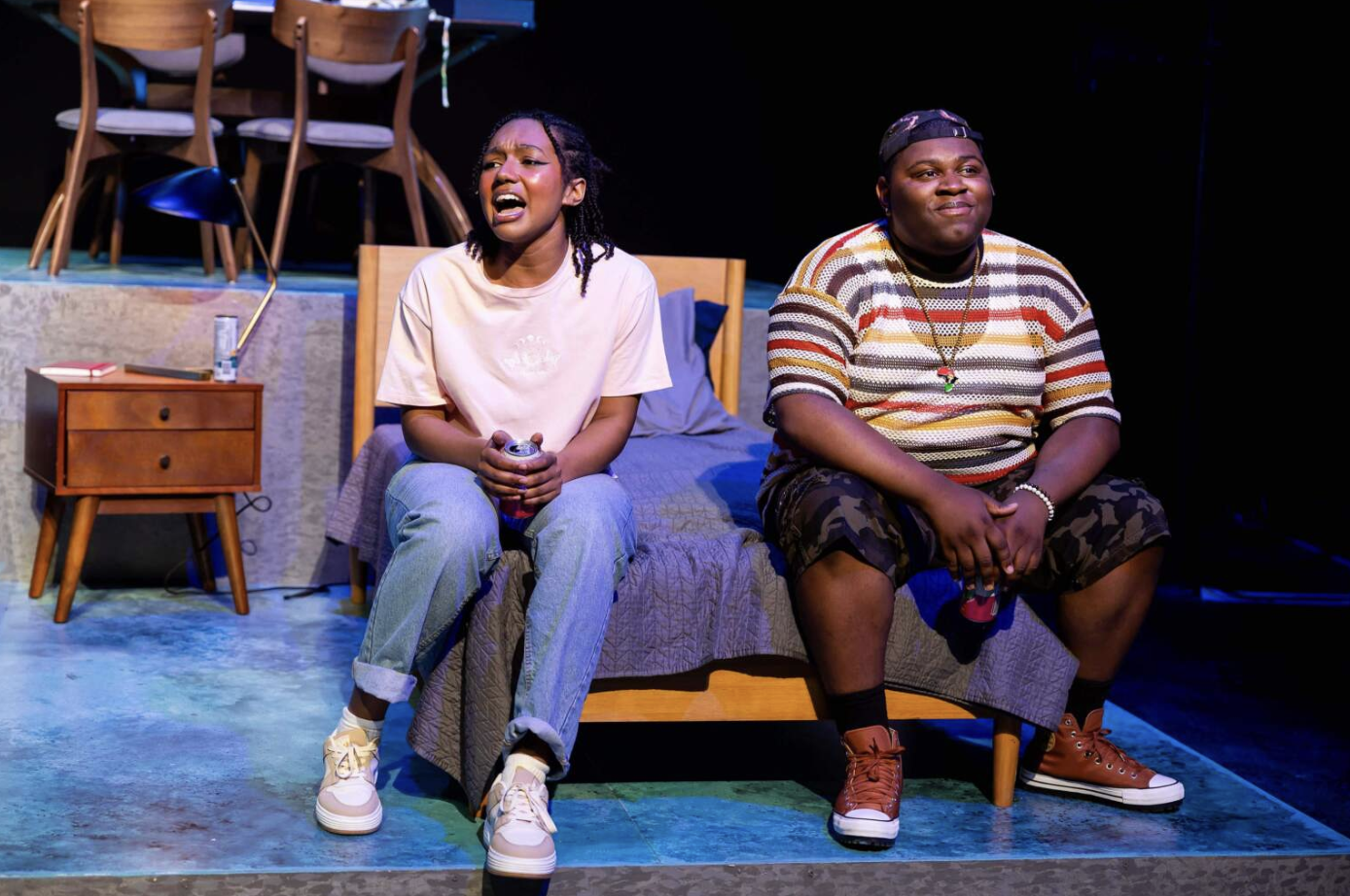 Two people sitting on a bed, one singing or talking passionately and the other smiling, in a stage play setting.