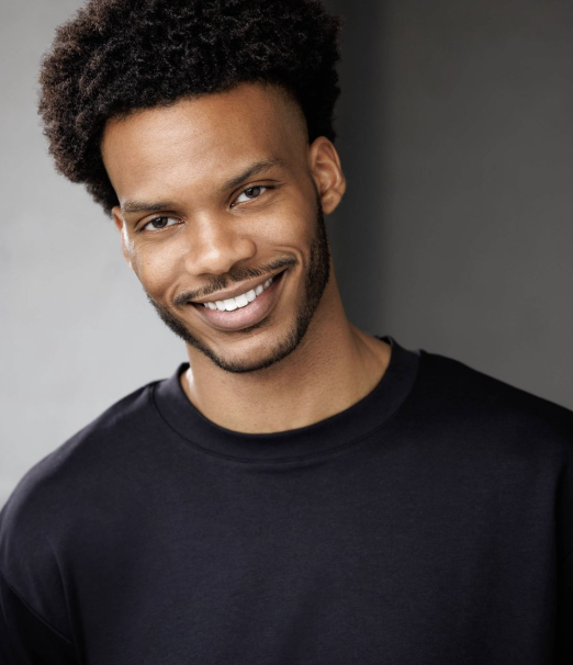 Portrait of a young man with curly hair and a beard smiling while wearing a black shirt.